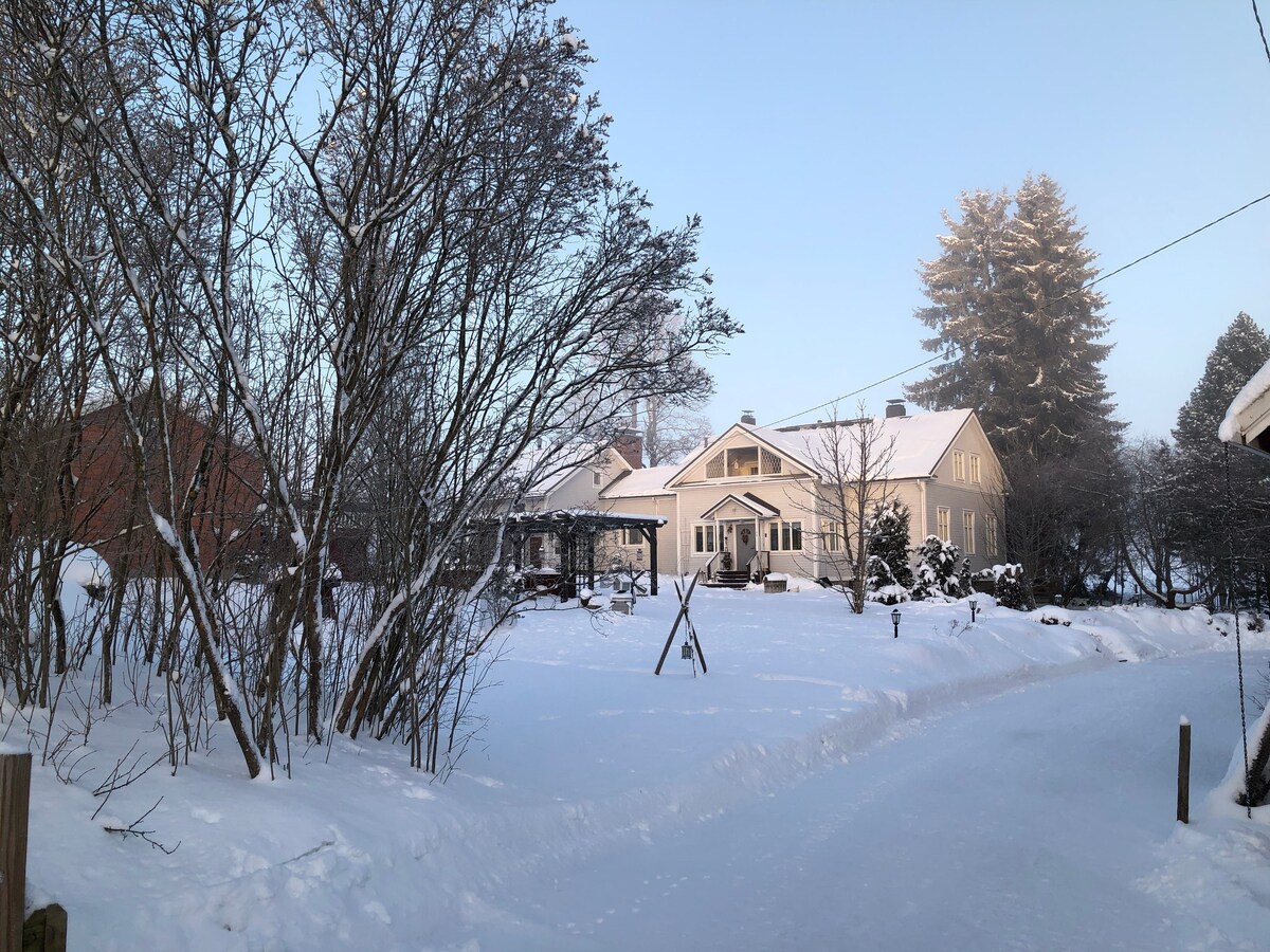A serene winter scene depicts a traditional countryside house surrounded by snow-covered landscape. Bare trees line the path leading to the home, with a gazebo visible on the property. The structure features light-colored walls and a sloped roof, contrasted against the white snow.