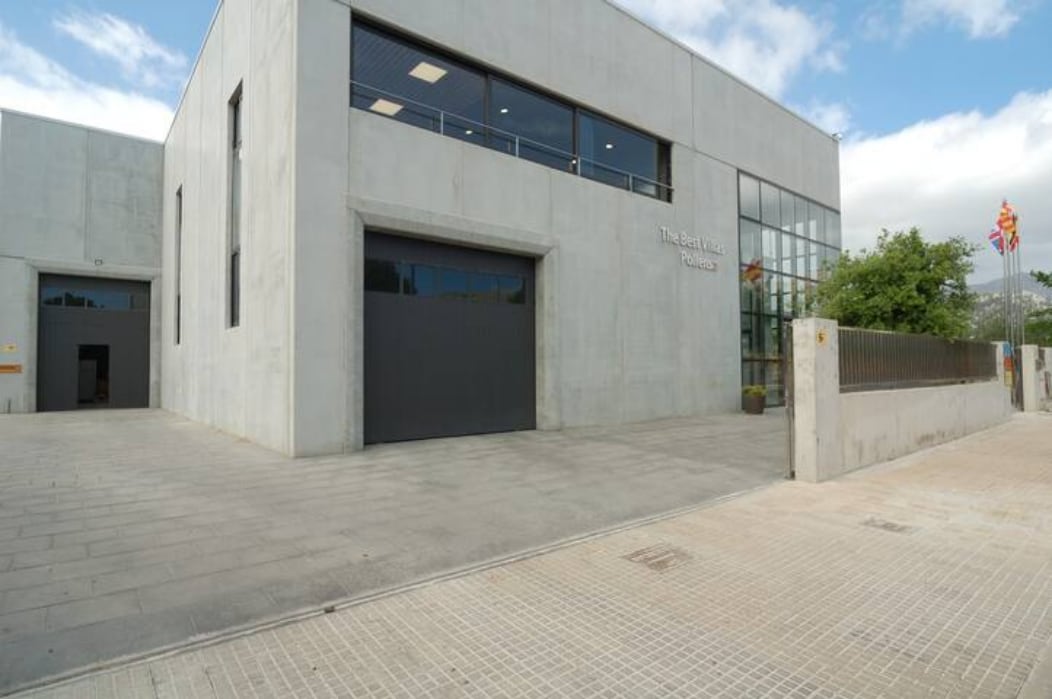 The exterior of the villa features a modern concrete facade with large windows allowing ample natural light. The entrance is marked by a wide, paved area leading to the main door. Greenery is visible alongside the building, enhancing the overall setting.
