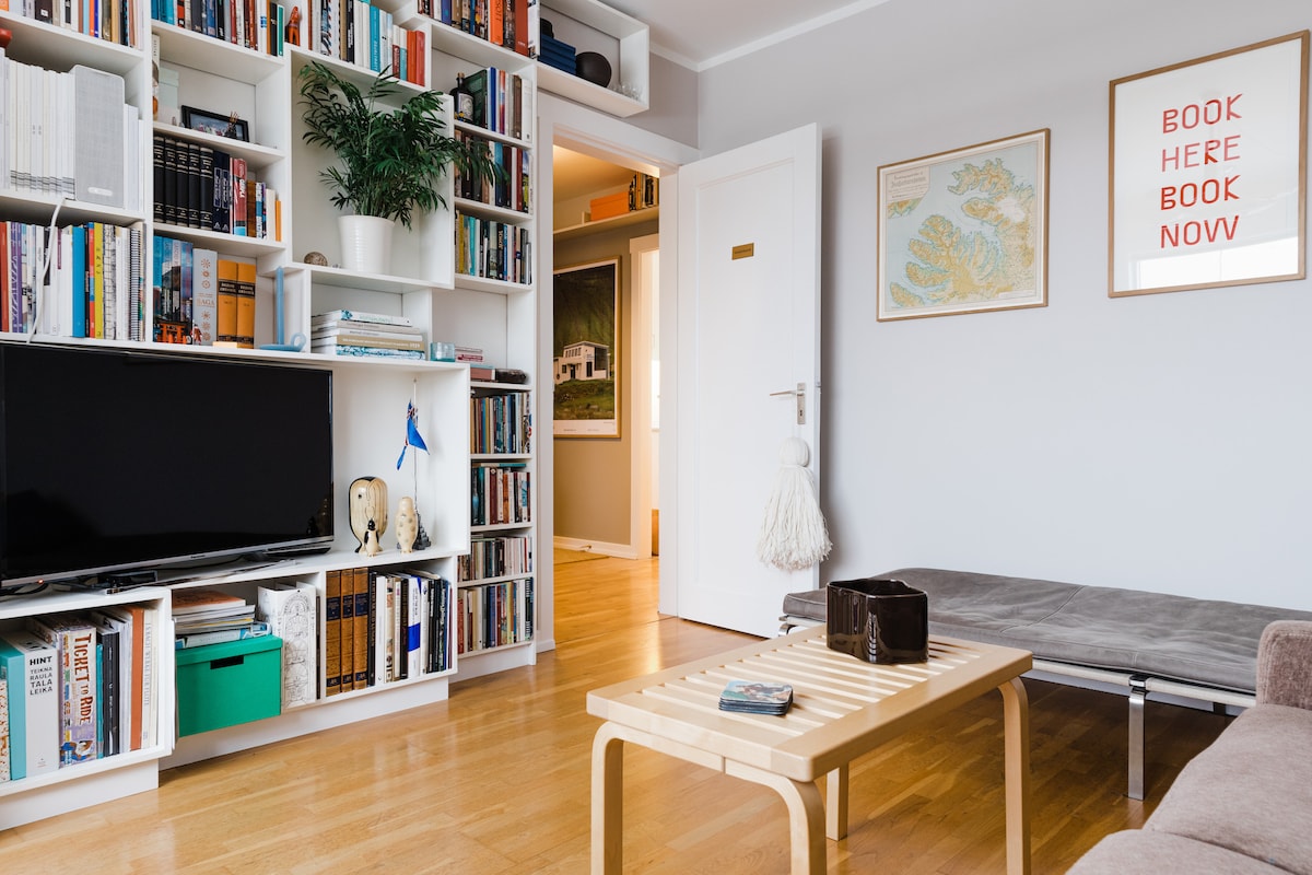 A bright living room is highlighted by a large shelf filled with books and decorative items. A flat-screen television is positioned on a modern stand, while a light wooden coffee table sits in the center. A comfortable sofa is visible, adding to the space's inviting nature.