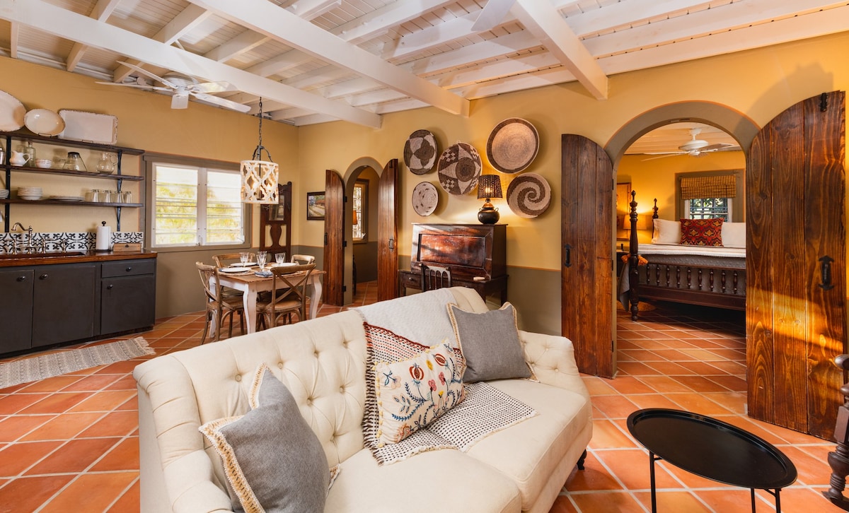 The living area features a comfortable white sofa adorned with patterned cushions. A dining table for four and a wooden cabinet are visible, along with warm terracotta tiles underfoot. Arched doorways lead to a cozy bedroom space, enhancing the open floor plan.