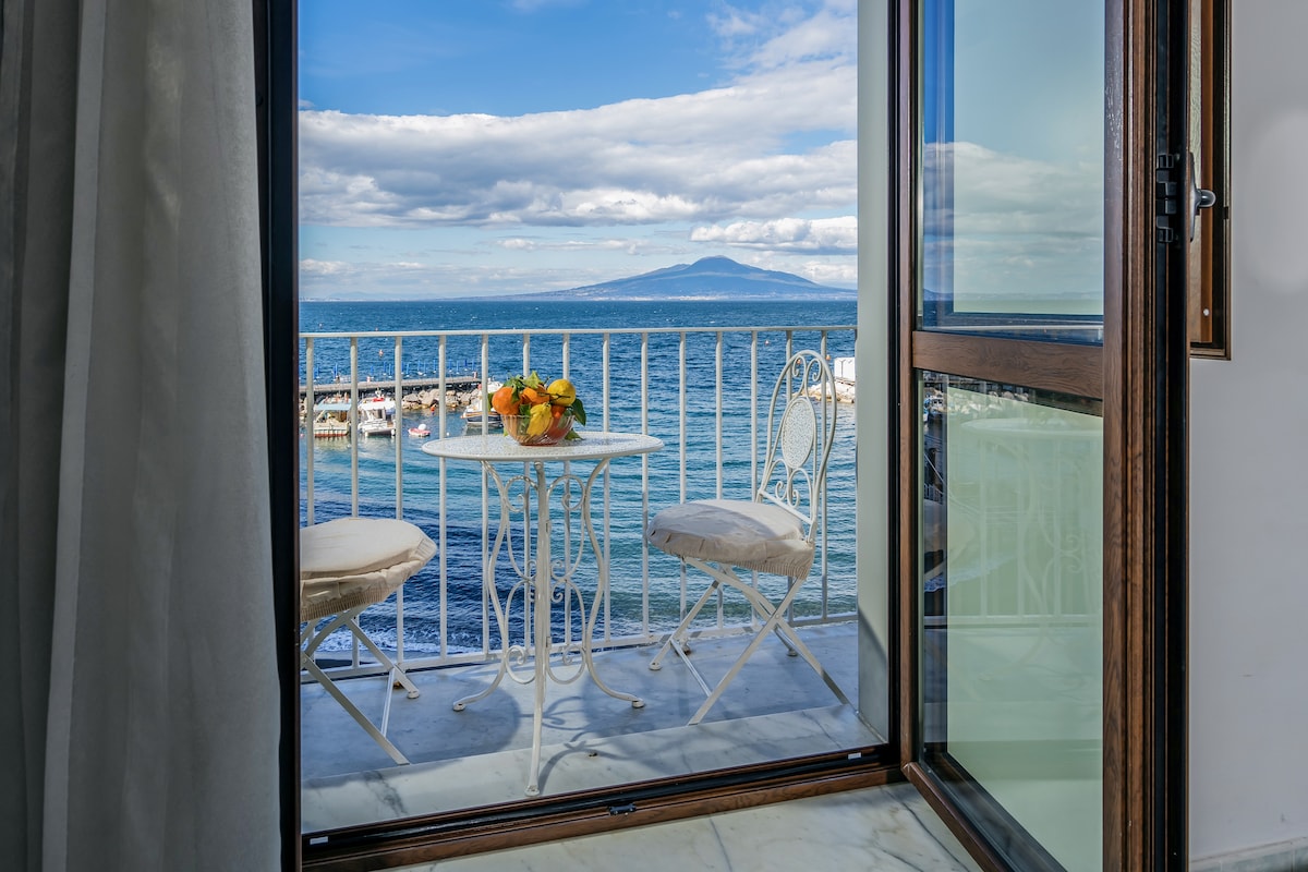 A small balcony features a round table and two chairs, overlooking the sea. The view includes the coastline and Mount Vesuvius under a partly cloudy sky. The glass doors provide access to the balcony, enhancing the bright and airy atmosphere.