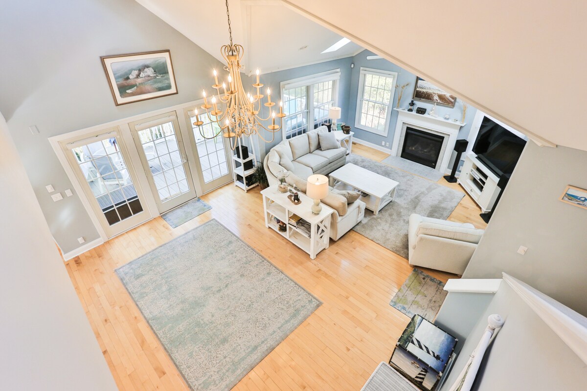 An open living area is captured from above, showcasing a light-filled space with large windows and wood flooring. A comfortable sectional sofa faces a coffee table, while a chandelier adds elegance. The room features welcoming light blue walls and a cozy rug.