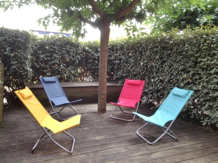 A private wooden terrace is surrounded by lush green hedges, featuring four reclining lounge chairs in yellow, blue, pink, and turquoise. The area is shaded by a tree, providing a serene space for relaxation.