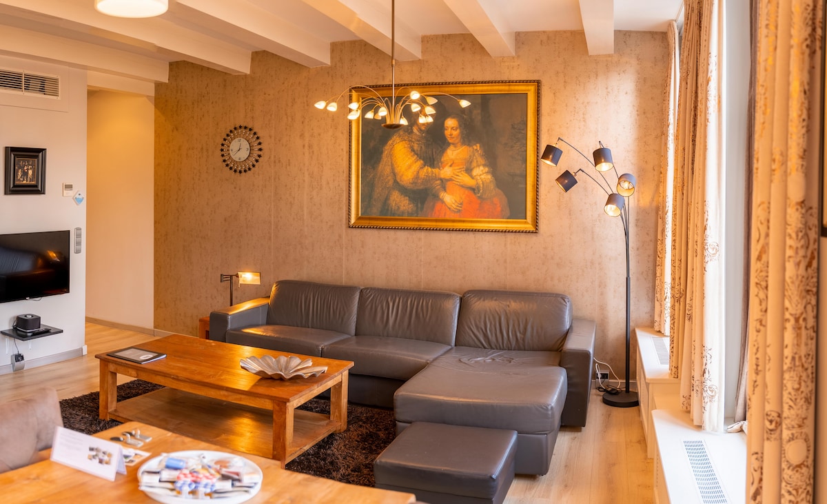 A spacious living area features a leather lounge sofa and a wooden coffee table, accompanied by a floor lamp and a wall clock. A large artwork hangs prominently above the sofa, while large windows allow natural light to fill the room, complemented by soft curtains.