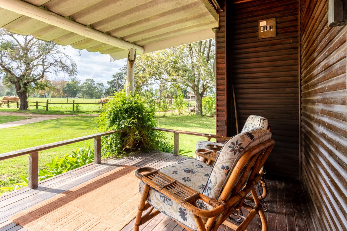 A shaded veranda is populated with two wooden chairs featuring patterned cushions. The view reveals a grassy area with trees and a fence in the background, creating a peaceful and open atmosphere.
