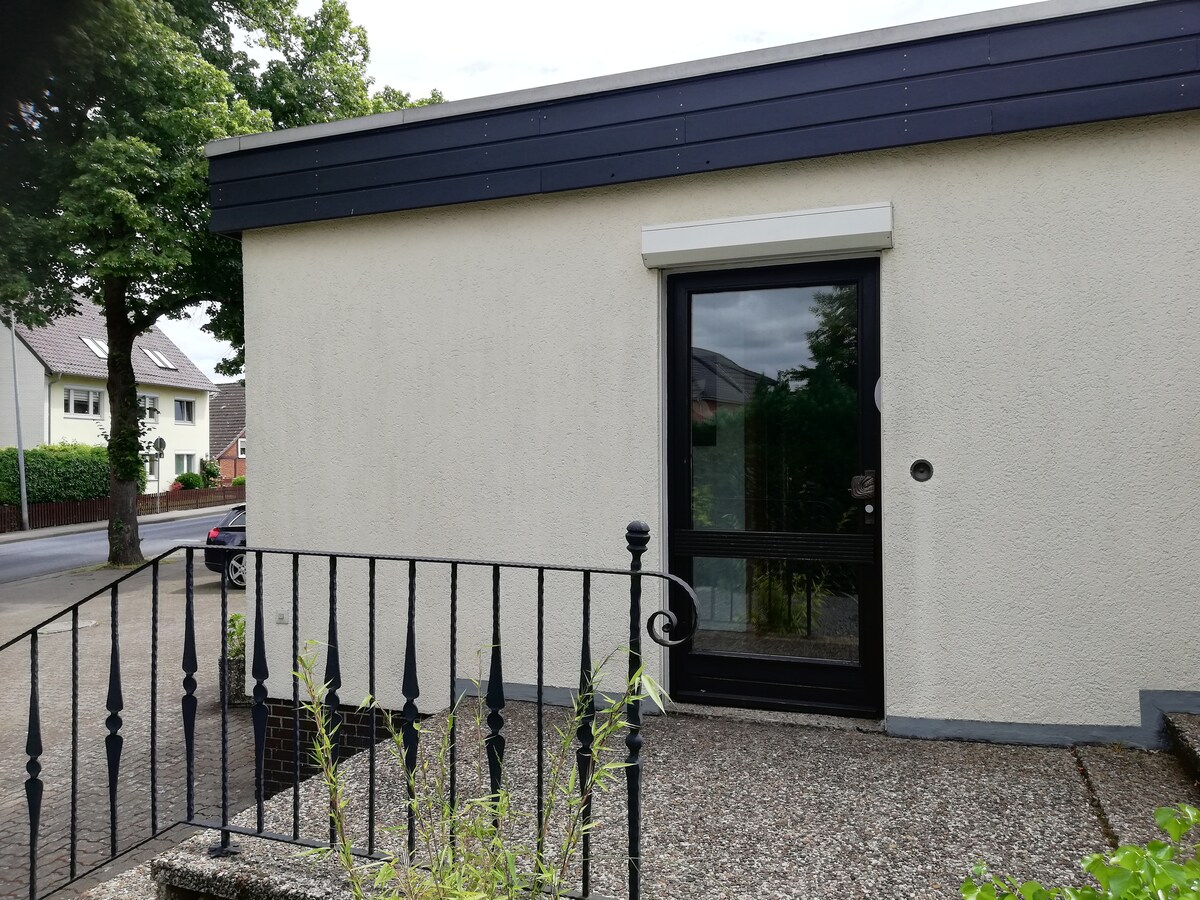 The image displays the entrance of the apartment, featuring a smooth surface with a dark door framed by light-colored walls. A railing borders a few steps leading up to the entrance, providing access to the inviting space.