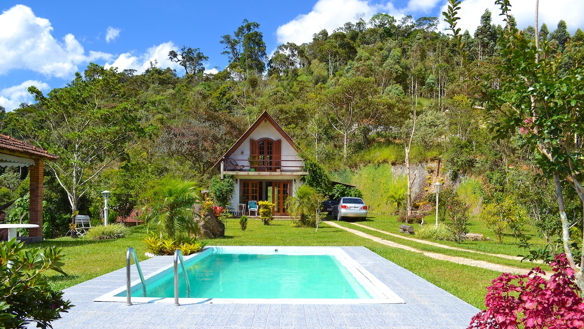A cozy chalet is set against a backdrop of lush greenery and mountains. A clear pool is positioned in the foreground, surrounded by a well-maintained lawn. Flowers and plants decorate the area, enhancing the serene outdoor space.
