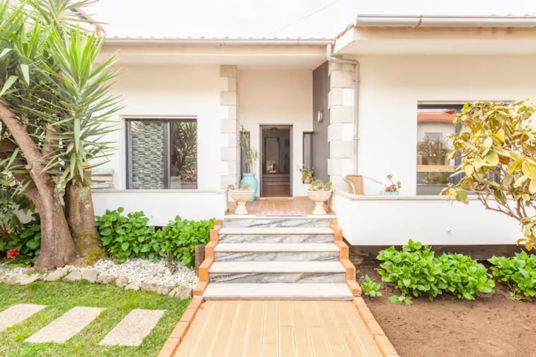 A welcoming entrance features a series of marble steps leading to the front door, framed by lush greenery and decorative plants. Large windows allow natural light to enter, highlighting the clean lines and open space of the home's facade.