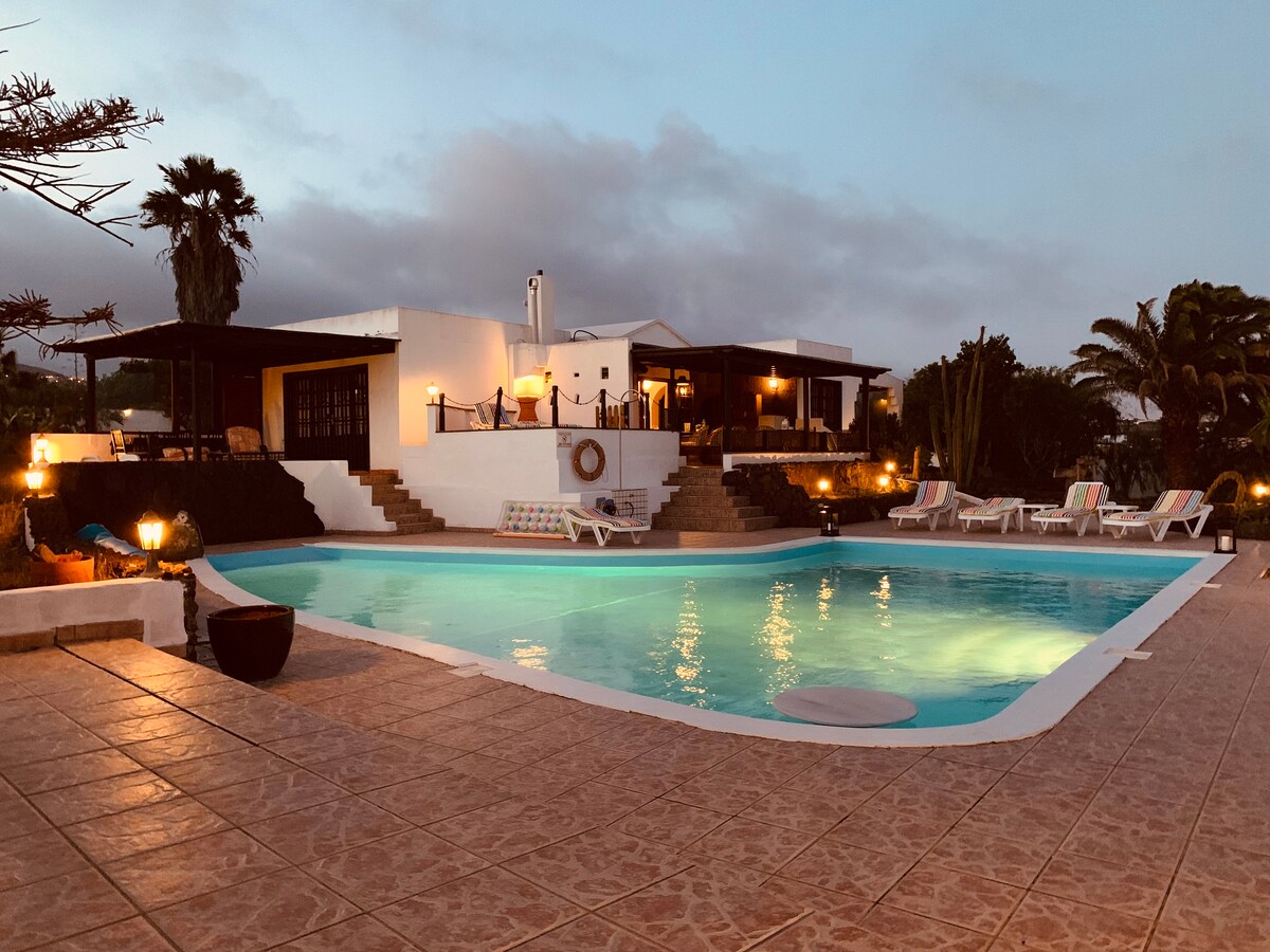A spacious outdoor area features a large L-shaped heated swimming pool, surrounded by sun loungers and tropical plants. The villa is visible in the background, with warm lighting enhancing the tranquil ambiance as dusk settles in.