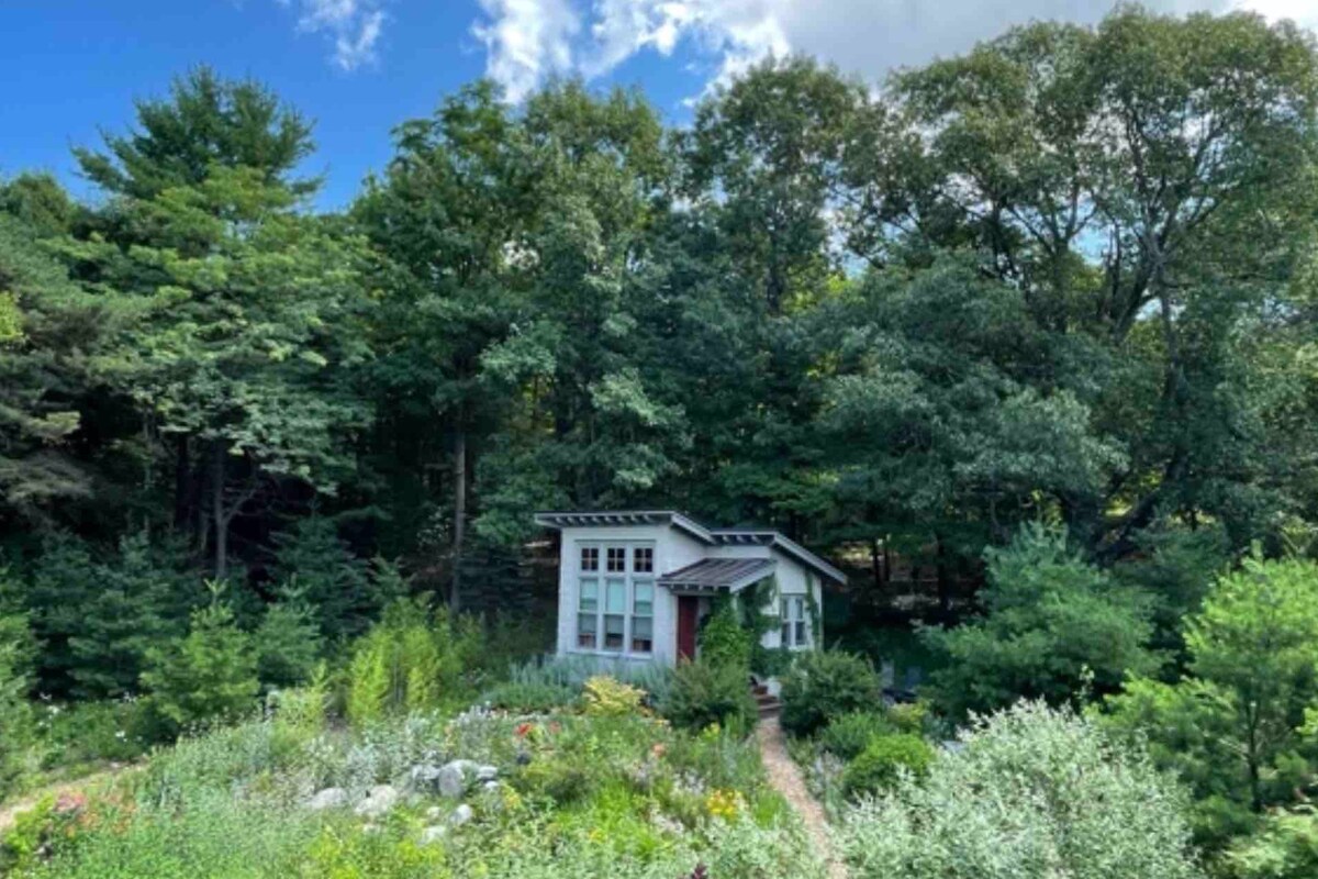 A small guesthouse is nestled among lush greenery and trees, surrounded by vibrant gardens. A winding stone pathway leads to the entrance, framed by blooming plants. The clear sky above adds a bright touch to the natural setting.