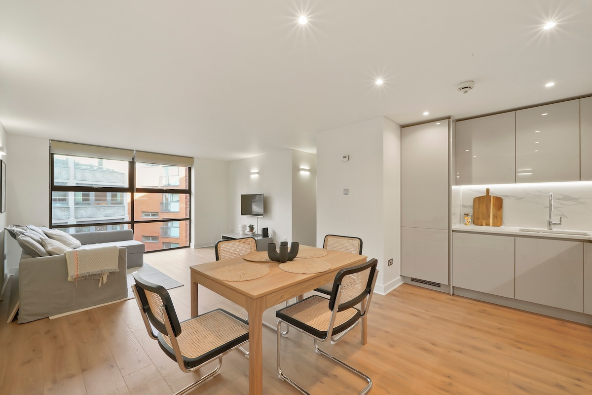 An open-plan living area is displayed, featuring a glass dining table surrounded by four chairs. A modern kitchen is positioned in the background, with sleek cabinetry and a countertop. Natural light enters through large windows, illuminating the light wood flooring and a comfortable sofa in the corner.