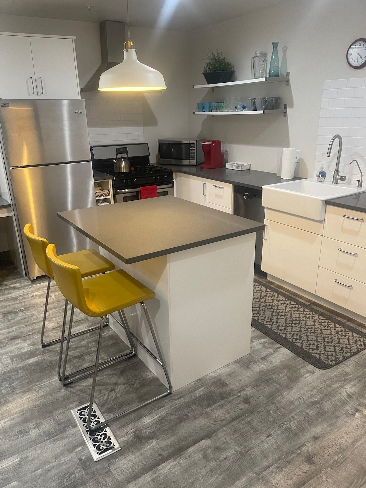 The kitchen area features modern appliances, including a stainless steel refrigerator and a microwave. A central island with two yellow bar stools provides a casual dining option. Sleek cabinetry and open shelving hold various kitchen items, with a farmhouse-style sink positioned against the wall.