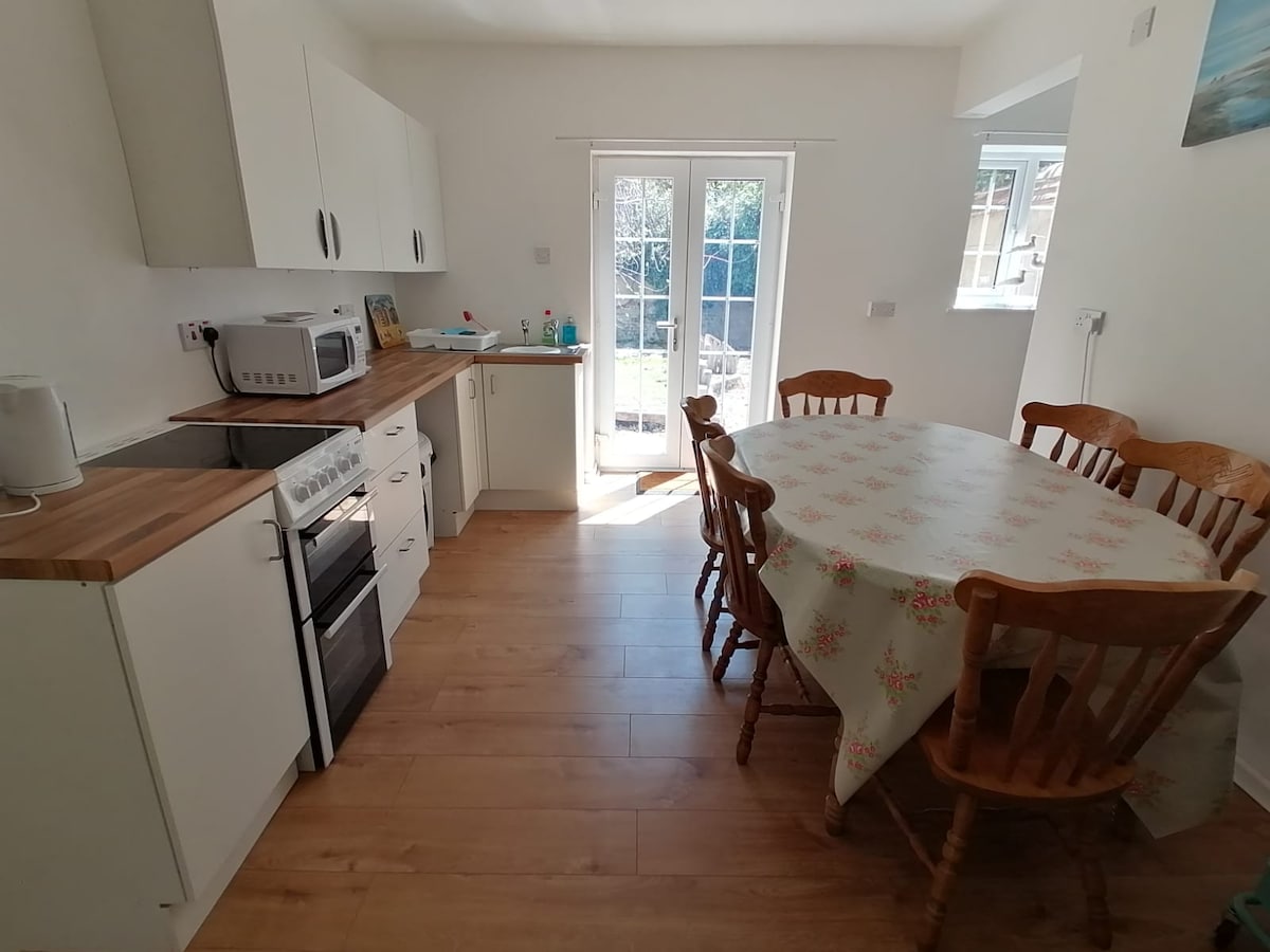 A bright kitchen area features white cabinetry and a wooden dining table surrounded by six chairs. A stove and microwave are visible on the countertop. Natural light enters through a set of French doors that lead to the outside space.