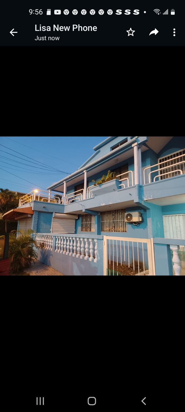 A two-story building with a vibrant blue exterior features a series of balconies adorned with plants. The front is framed by a white railing, and large windows provide ample natural light. The scene is complemented by a clear sky, enhancing the inviting atmosphere.
