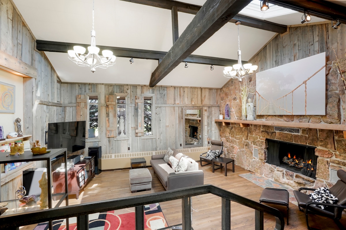 A spacious living area features wooden paneling and exposed beams, creating a warm ambiance. A stone fireplace is surrounded by comfortable seating, with a large television displayed on a wall. Natural light enters through multiple windows, enhancing the room's inviting feel.