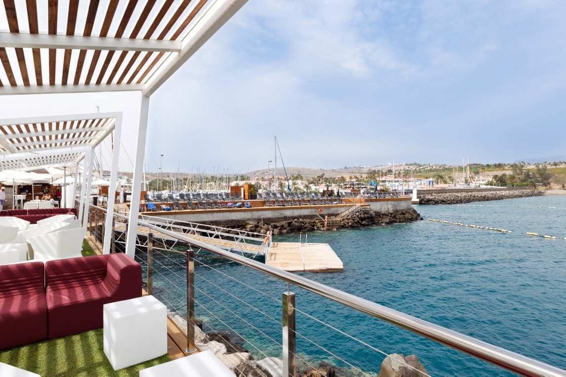 A waterfront terrace offers views of a marina with numerous boats docked. Comfortable seating arrangements in white and maroon are placed under a pergola, providing shade. The water reflects the sky, creating a serene atmosphere. A wooden boardwalk extends toward the marina.