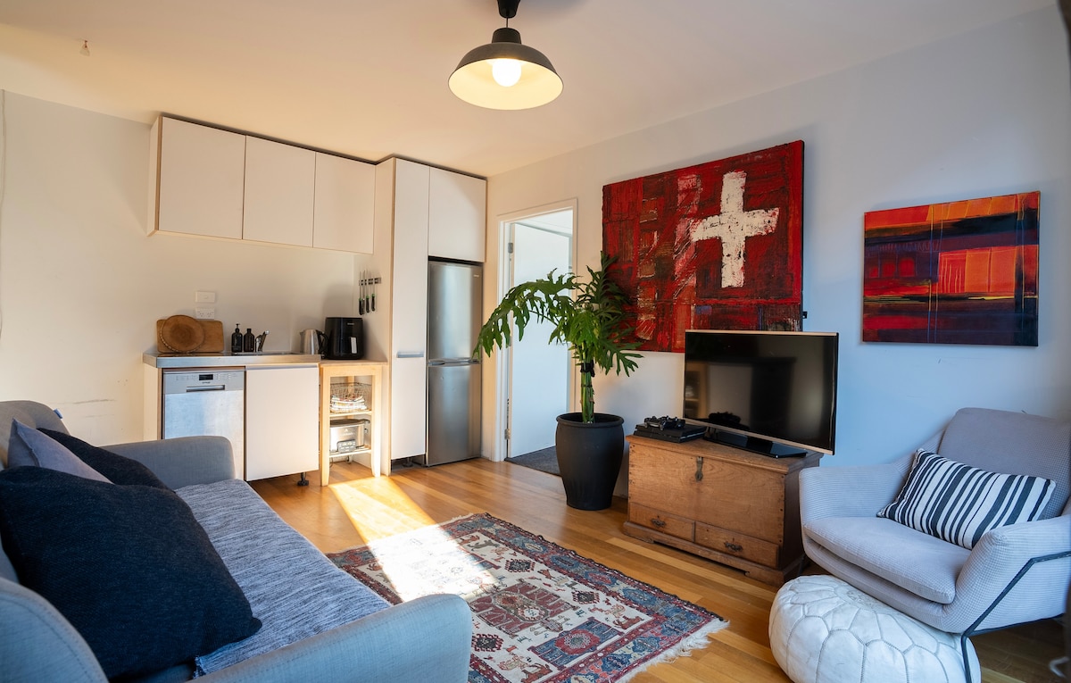 A bright living area features a comfortable sofa and a light-colored armchair beside a vintage-style storage chest. A flat-screen television is positioned on a low stand. A kitchenette with white cabinetry and a small dining area is visible. Colorful artwork adorns the walls.