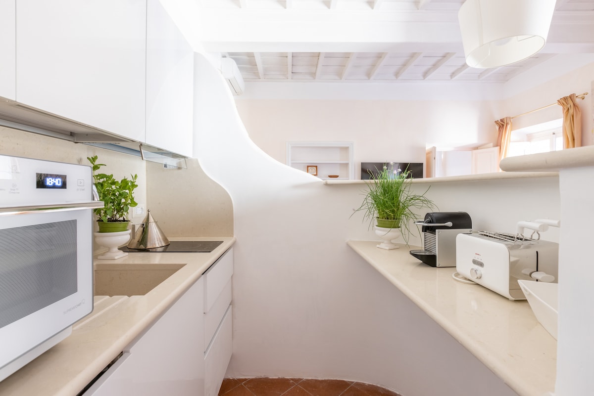 The kitchenette is designed with modern white cabinetry and a light countertop. Appliances such as a microwave, toaster, and coffee maker are arranged neatly. Plants are placed for a touch of greenery. Natural light is observed from nearby windows featuring soft curtains.