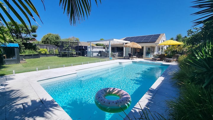 Sunny backyard with a swimming pool, lounge chairs, umbrellas, a trampoline, and a house with solar panels and a patio area