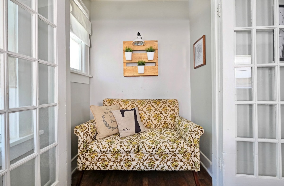 A cozy seating area features a patterned couch adorned with decorative pillows. Above, a wooden shelf holds plants, adding a touch of greenery to the space. Bright natural light filters in through the surrounding glass-paneled doors.