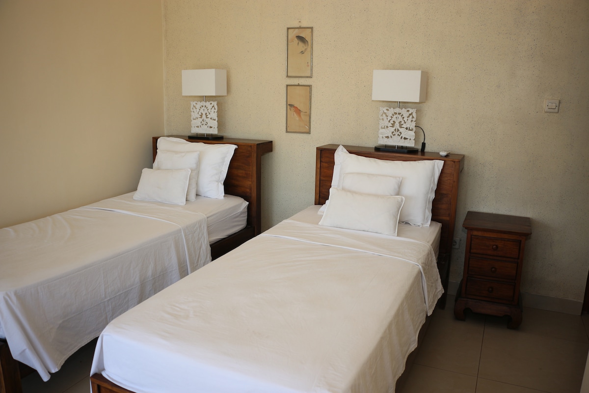 A well-lit room features two single beds with crisp white linens and multiple pillows. Each bed is positioned next to wooden nightstands, which hold modern lamps. Soft artwork hangs on the wall above the nightstands, adding a subtle decorative touch.