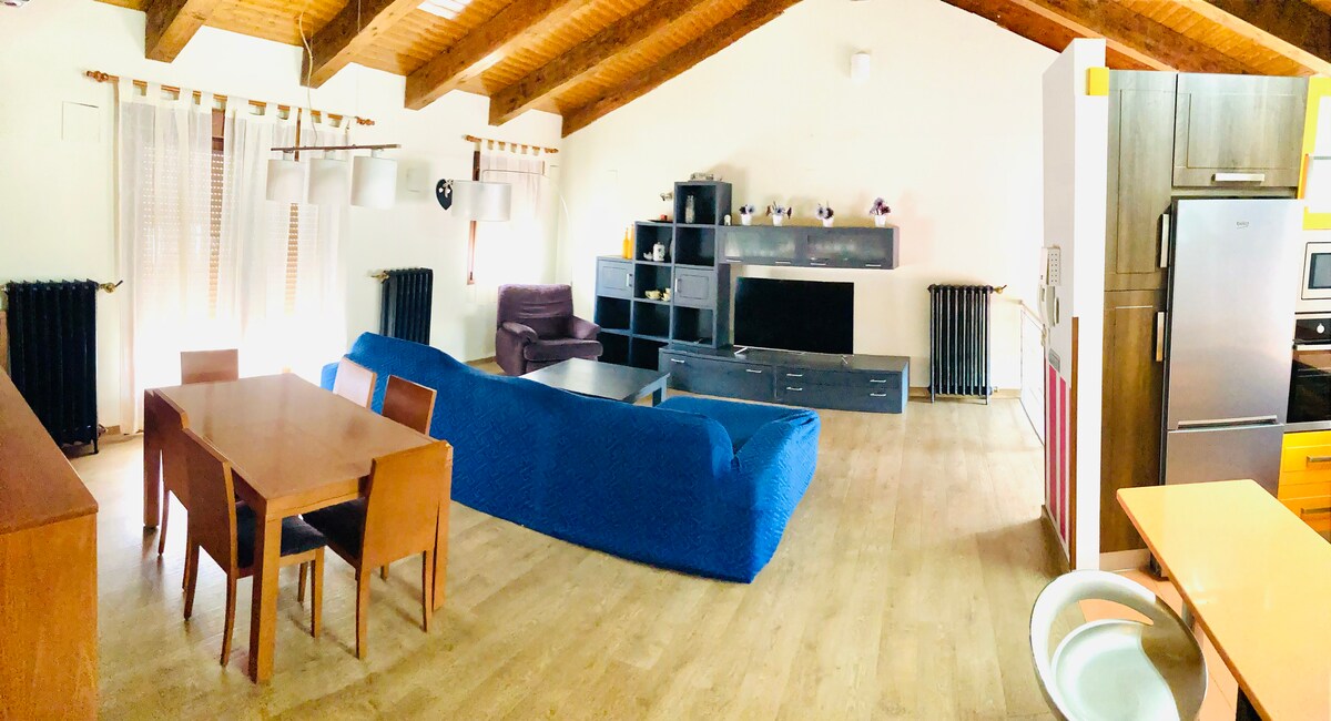 An open living area features a large blue sofa, a dining table with wooden chairs, and a contemporary entertainment unit. Soft natural light filters through sheer curtains, illuminating the warm wooden beams overhead. A cozy armchair is situated nearby, adding to the space's comfort.