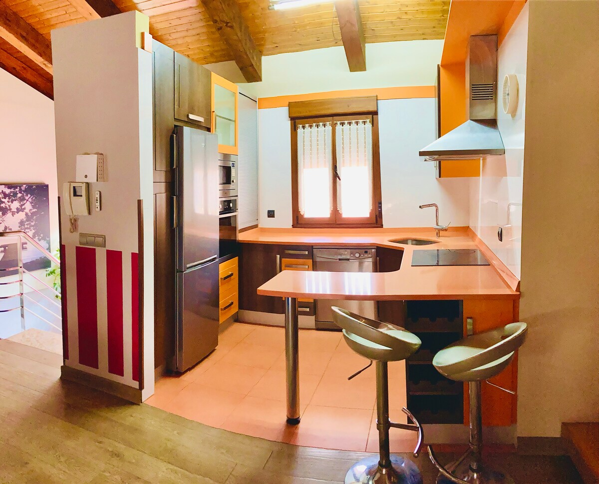 The kitchen features a modern setup with wooden beams overhead. Stainless steel appliances are positioned alongside a vibrant orange countertop. A window with vertical blinds allows natural light to fill the space. Two high-backed stools are positioned at the counter for seating.