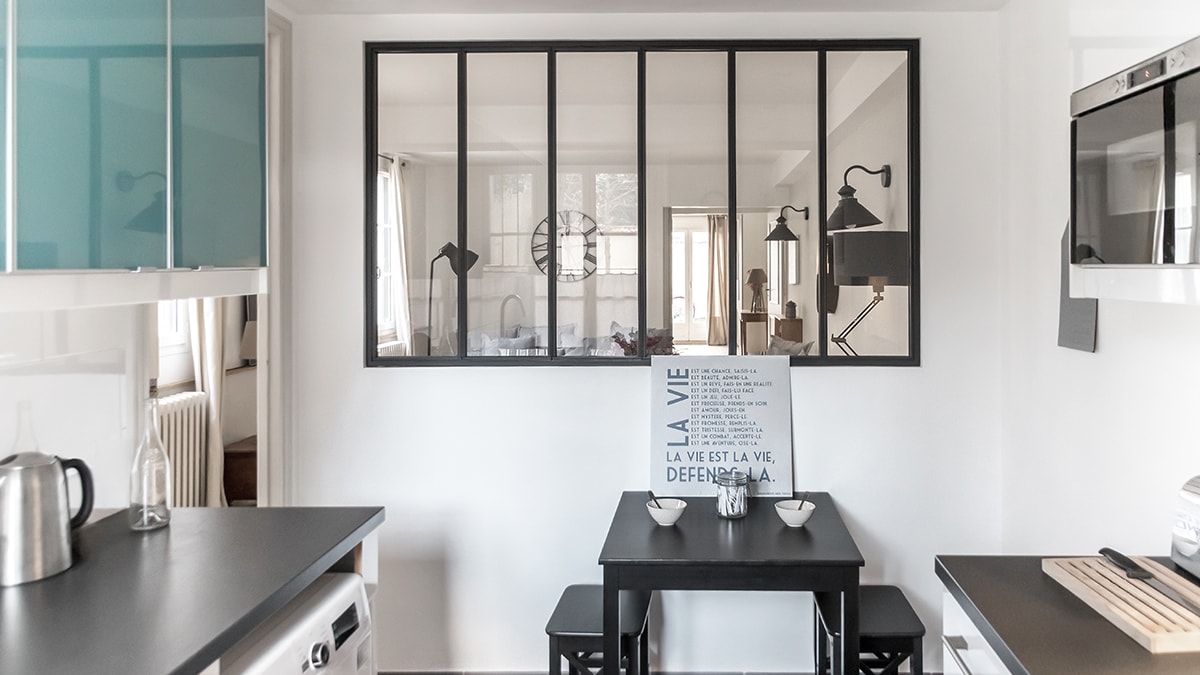 The contemporary kitchen area features a sleek black table set with two bowls. A glass partition allows light to flow between the spaces, while the minimalist design is complemented by light-colored cabinetry and stainless steel appliances. Natural light enters through nearby windows.