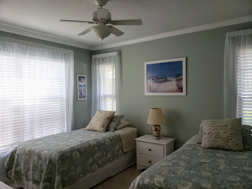 A serene bedroom features two twin beds with light floral bedding and decorative pillows. Natural light filters through sheer white curtains, highlighting a calm atmosphere. A bedside lamp rests on a white nightstand, and a wall art piece depicting a boat adds a subtle nautical touch.