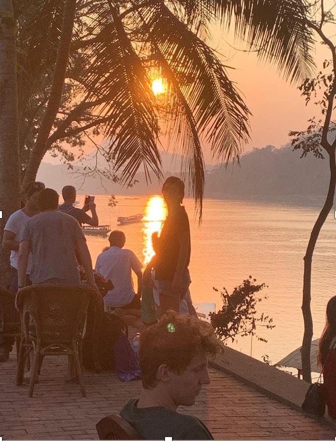 People are gathered by the river at sunset, with warm golden light reflecting off the water. Silhouettes of trees frame the scene, creating a peaceful atmosphere. Several individuals are seated and standing, enjoying the view of the Mekong River and distant hills.