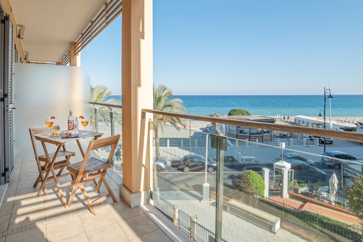A private balcony is presented, featuring a small wooden table set with drinks and two chairs. The view overlooks a calm beach and the sea, with palm trees lining the beachfront. Natural light illuminates the space, enhancing the coastal atmosphere.