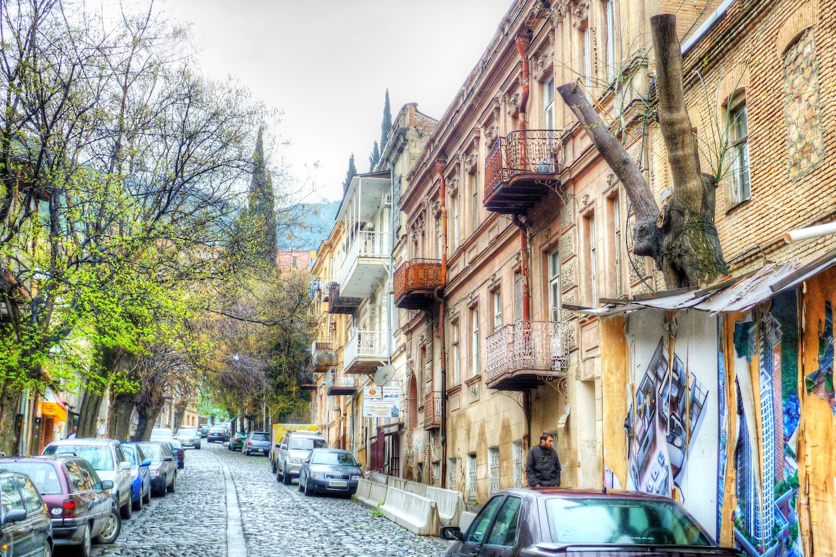 A cobblestone street lined with traditional Georgian buildings, showcasing balconies and artistic murals. Trees with budding leaves provide a touch of greenery, while parked cars are seen along both sides of the street. The serene atmosphere invites exploration of the local charm.