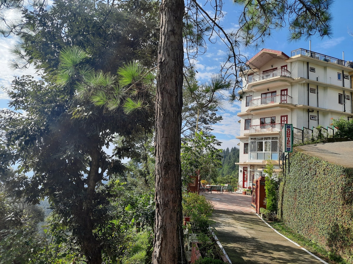 A multi-story villa is positioned on a hillside, surrounded by lush greenery. Tall trees frame the scene, offering shade. A pathway winds up to the villa, with seating areas visible in the distance. The sky above is clear with scattered clouds.