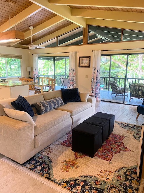 A spacious living area is illuminated by natural light from large windows, revealing a comfortable sofa adorned with navy blue accent pillows. A decorative rug anchors the space, with a dining area and wrap-around deck visible in the background, creating a seamless indoor-outdoor flow.