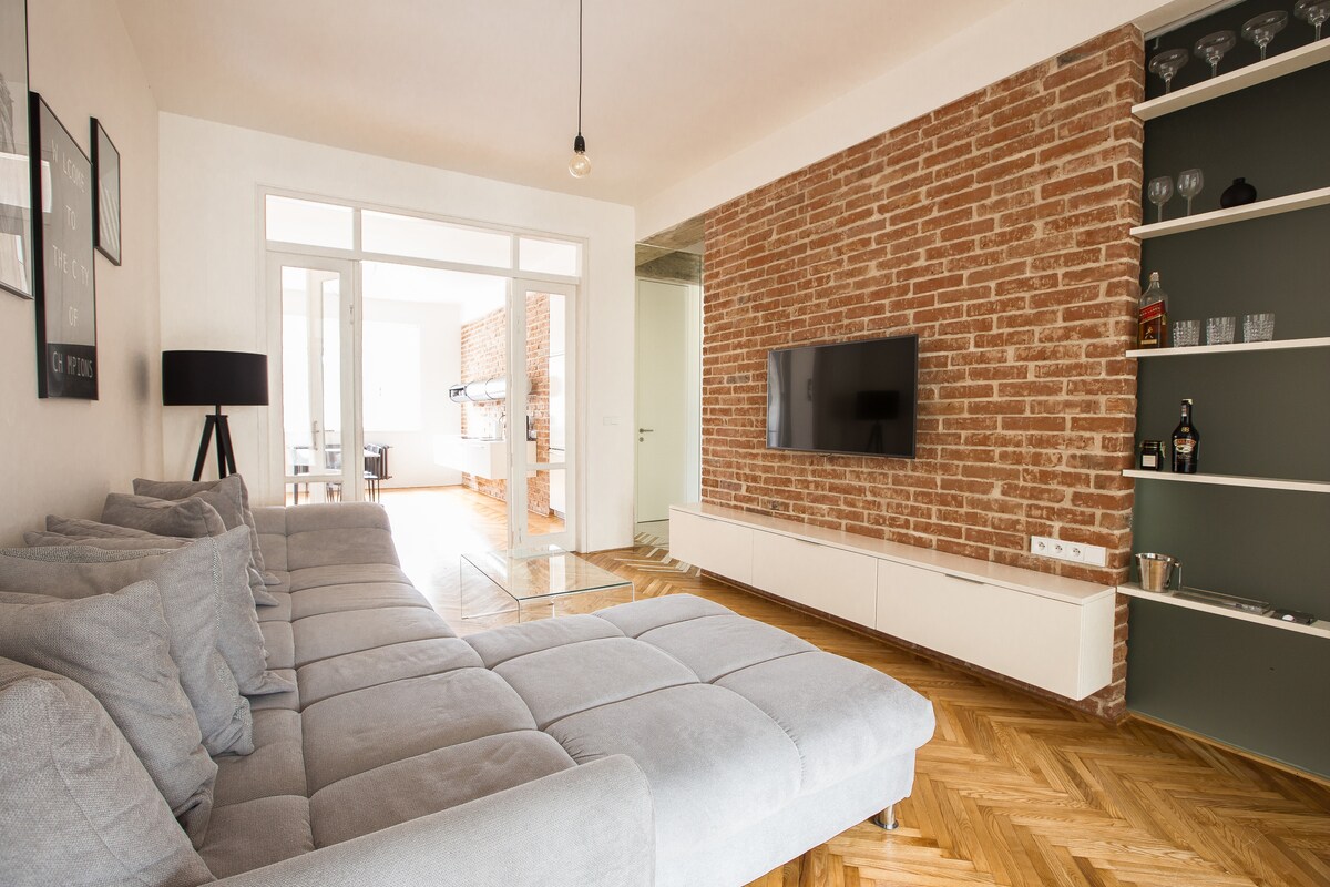 A spacious living area is presented with a comfortable gray sofa positioned against a textured brick wall. A flat-screen TV is mounted on a white cabinet, while decorative glassware is displayed on a nearby shelf. Natural light flows through glass doors leading to a balcony.