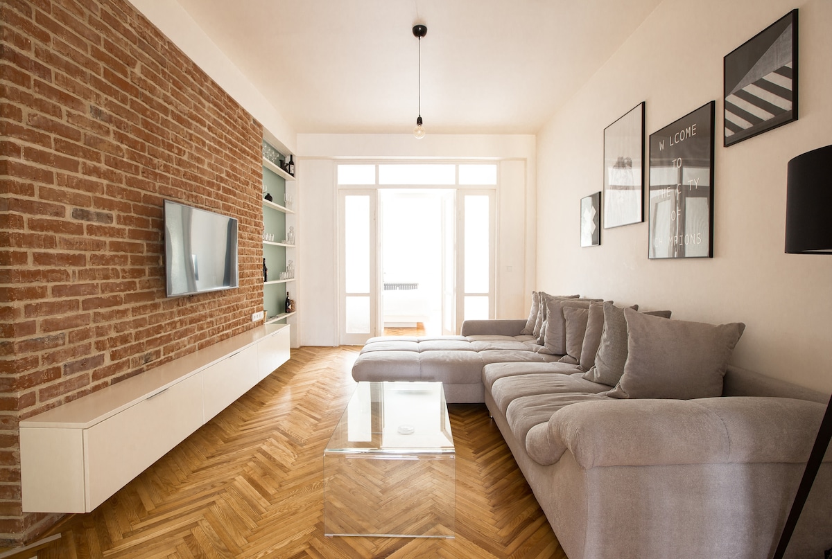 A spacious living area features a large sectional sofa in a neutral tone, complemented by a glass coffee table. A minimalist media unit houses a television mounted on a textured brick wall. Natural light fills the room through large glass doors leading to the balcony.