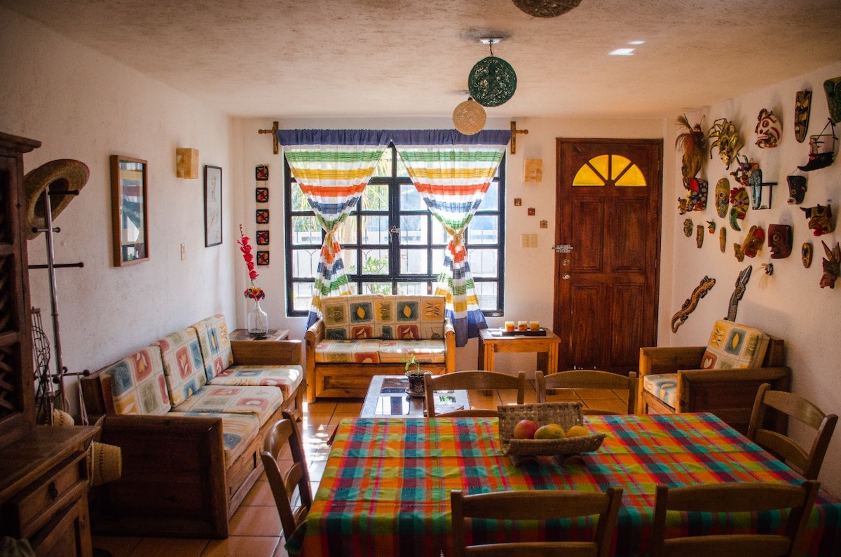 The living area is filled with natural light, featuring a colorful checkered tablecloth on a dining table surrounded by wooden chairs. Cozy seating is arranged with patterned cushions near large windows adorned with striped curtains. Walls display various traditional Mexican masks, enhancing the cultural ambiance.