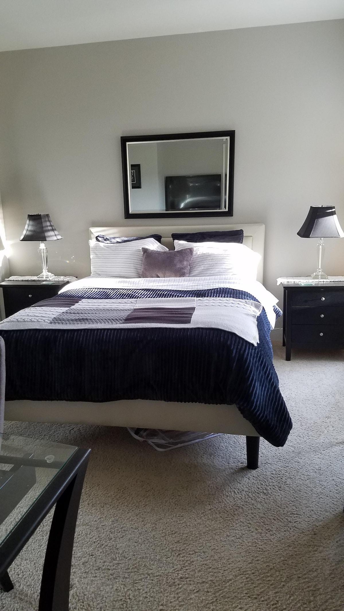 A well-lit bedroom contains a compact bed with a layered bedding arrangement featuring various shades of purple and navy. Two bedside tables with lamps are situated on either side, and a large mirror is positioned above the bed. A flat-screen TV is visible on the opposite wall.