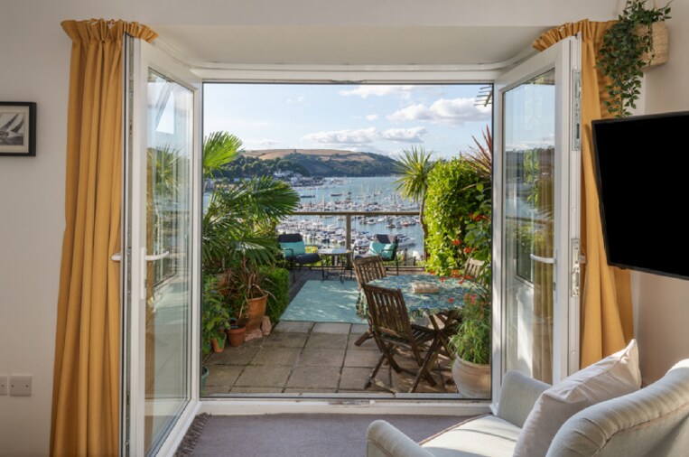 Double French doors open to reveal a private terrace adorned with potted plants and outdoor dining furniture. The stunning river views showcase the river Dart and nearby hills, enhancing the tranquil ambiance of the studio.