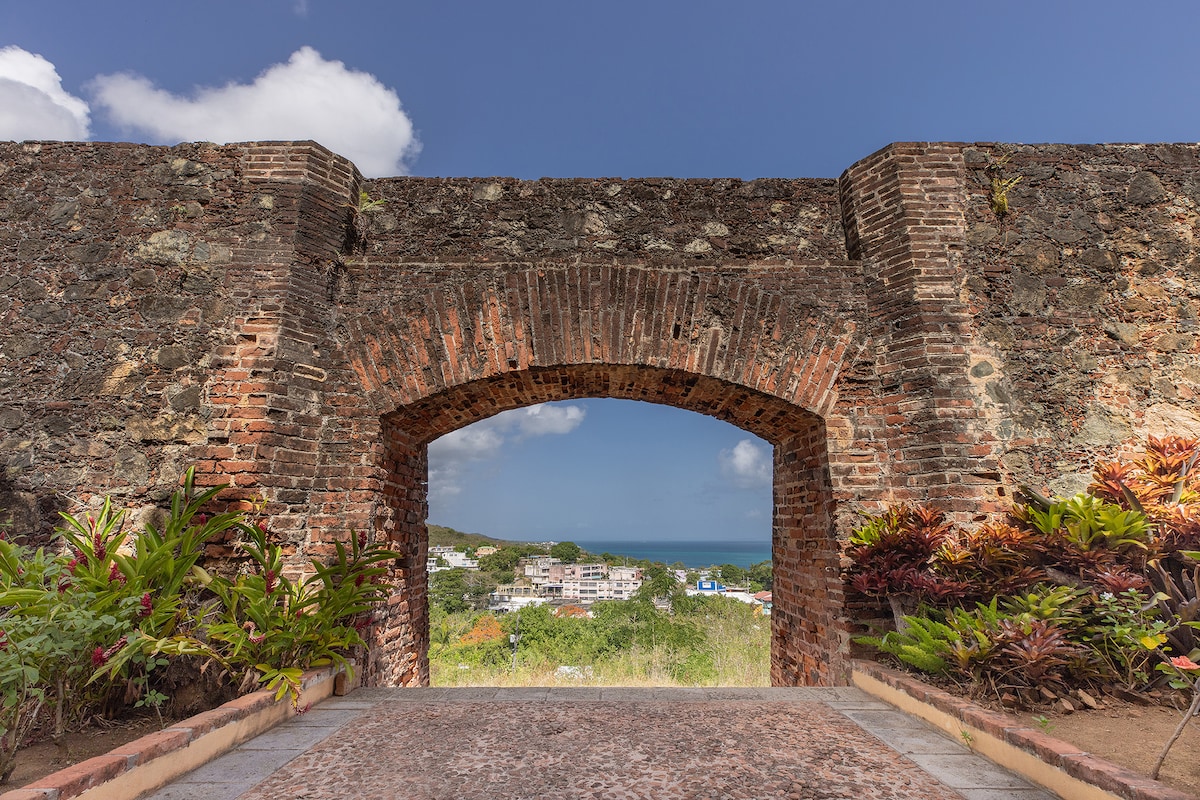 An arched brick doorway frames a view of the vibrant town and coastline beyond. Lush greens and colorful plants line the pathway leading up to the arch, creating a harmonious blend of natural beauty and historic architecture under a clear blue sky.