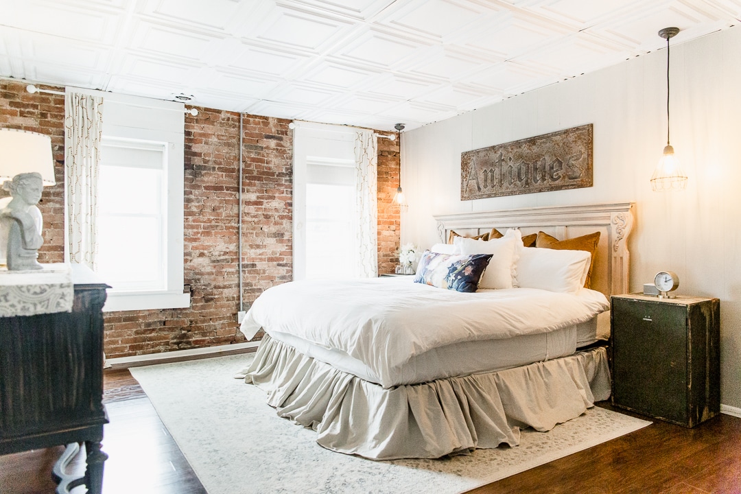 A spacious sleeping area features a large bed with a softly ruffled bed skirt. Exposed brick walls complement soft white linens and decorative pillows. Natural light streams in through two windows, while pendant lights add subtle illumination, creating a welcoming ambiance.