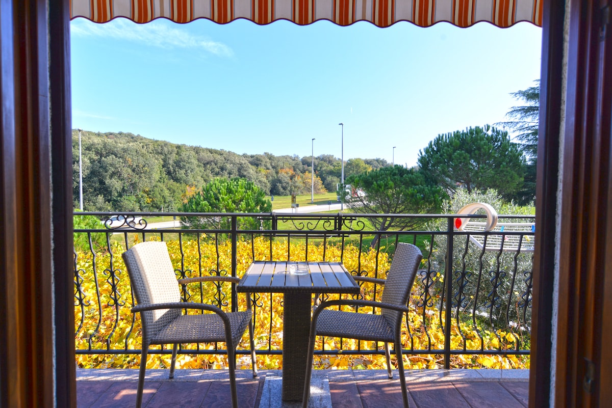 A private balcony is shown, featuring a small table and two comfortable chairs. Lush greenery and vibrant foliage are visible beyond the railing, complemented by a blue sky overhead and a striped awning offering shade.