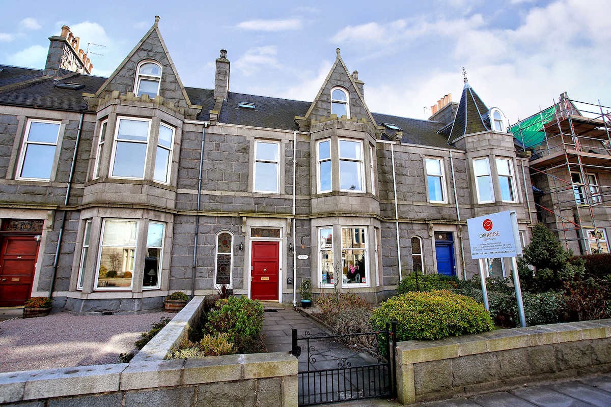 A charming stone-fronted building showcases distinctive architectural features, including gabled roofs and large windows. Colorful doors are visible at the entrance, adding character to the overall façade. A well-maintained garden area is seen in front, contributing to the welcoming appearance of the property.