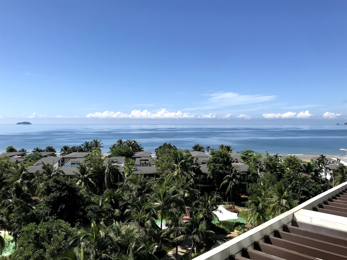 A panoramic view of the ocean is captured from an elevated balcony, showcasing the serene blue waters and a clear sky above. Lush greenery surrounds the resort buildings below, creating a tranquil and inviting atmosphere, complemented by distant islands visible on the horizon.