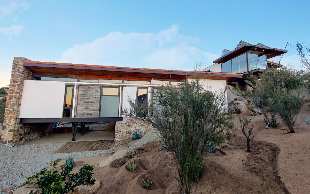 The architectural structure is integrated into the natural landscape, featuring a blend of stone and wooden elements. A spacious deck extends from the main building, with large windows providing views of the surroundings. The design reflects a harmony with nature.