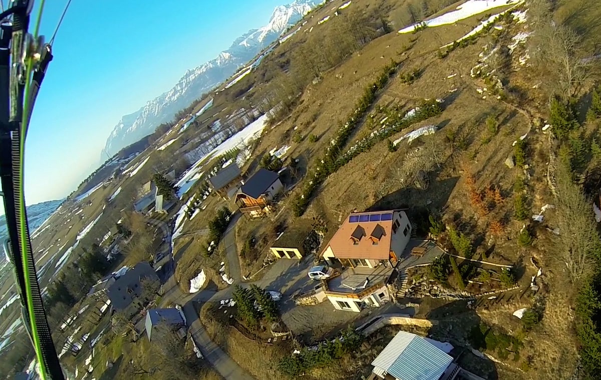 Aerial view reveals the charming village nestled among rolling hills and snow-capped mountains. The building features a red-tiled roof and is surrounded by winding paths and patches of greenery. Snow remains in shaded areas, contrasting with the earthy tones of the landscape.