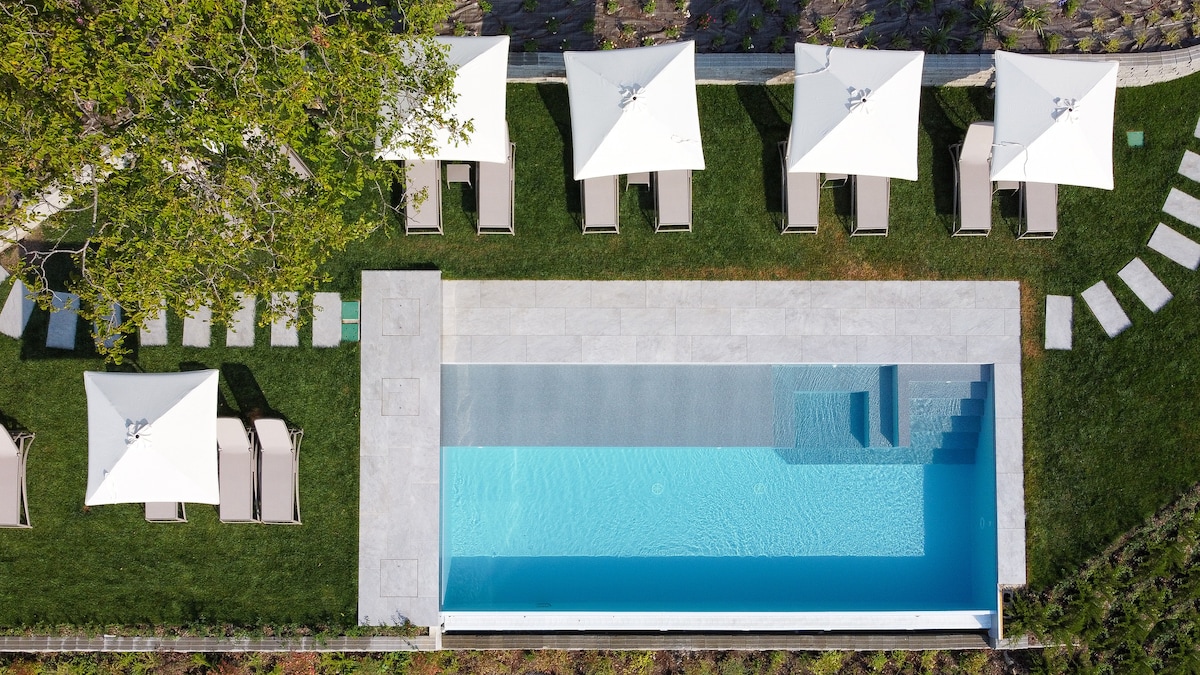 An aerial view of the infinity pool reveals tranquil turquoise water surrounded by a manicured lawn. Sun loungers with umbrellas provide shaded seating, and stone pathways lead to the pool area, making it an inviting space for relaxation.