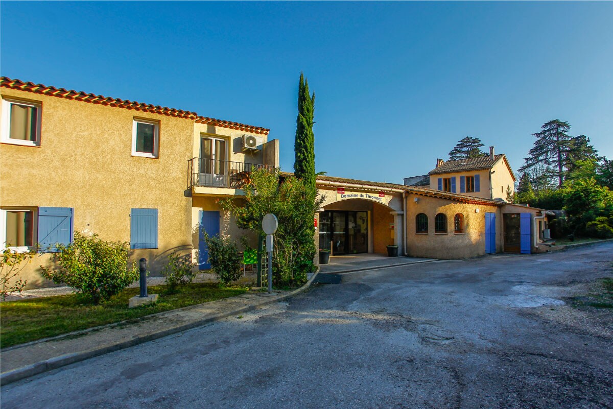 A charming residence featuring typical Provençal architecture, surrounded by greenery. Blue shutters adorn the windows, and an inviting entrance leads to the interior. The path is lined with shrubs, creating a welcoming atmosphere. The clear sky enhances the tranquil setting.