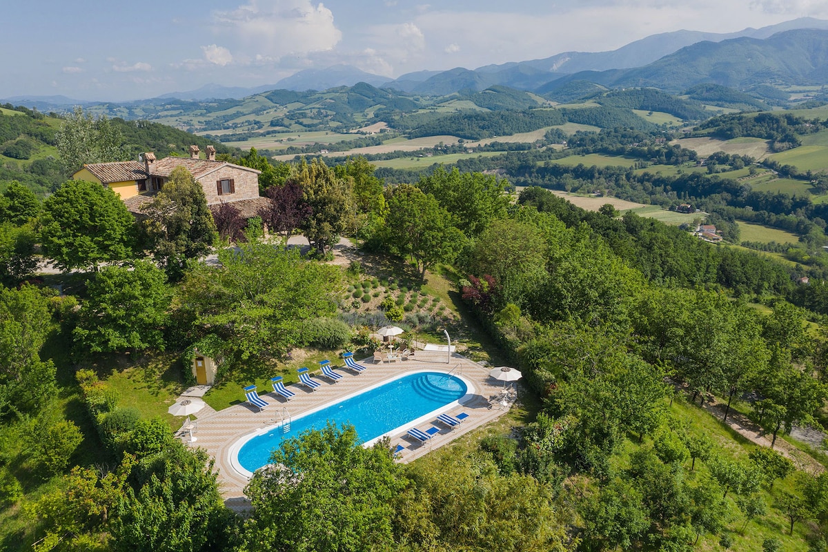 A spacious outdoor pool is surrounded by lush greenery and sun loungers. The property is nestled on a hillside with sweeping views of rolling hills and valleys. An inviting patio area and umbrellas offer shaded seating options, creating a relaxing environment.