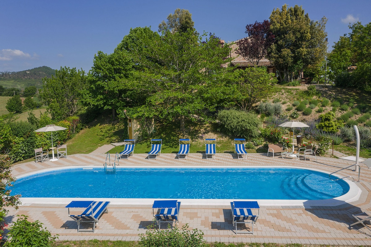 A spacious outdoor swimming pool is surrounded by sun loungers with blue-and-white stripes. Lush greenery, including trees and shrubs, enhances the landscape. A few umbrellas provide shade, while gentle hills are visible in the background, contributing to a serene setting.